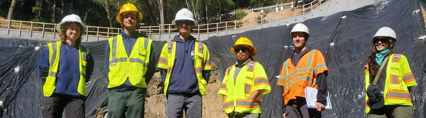Six people in vests and hard hats pose near a construction site