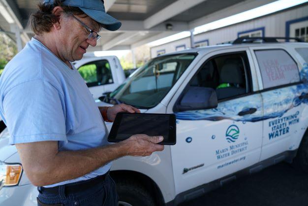A worker displays an electronic tablet in front of an SUV with Marin Water labeling.