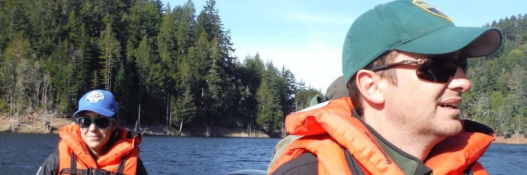 Two people, wearing hats and life jackets, ride in a boat on a Marin Water reservoir. Tree-lined hillsides span out in the background.