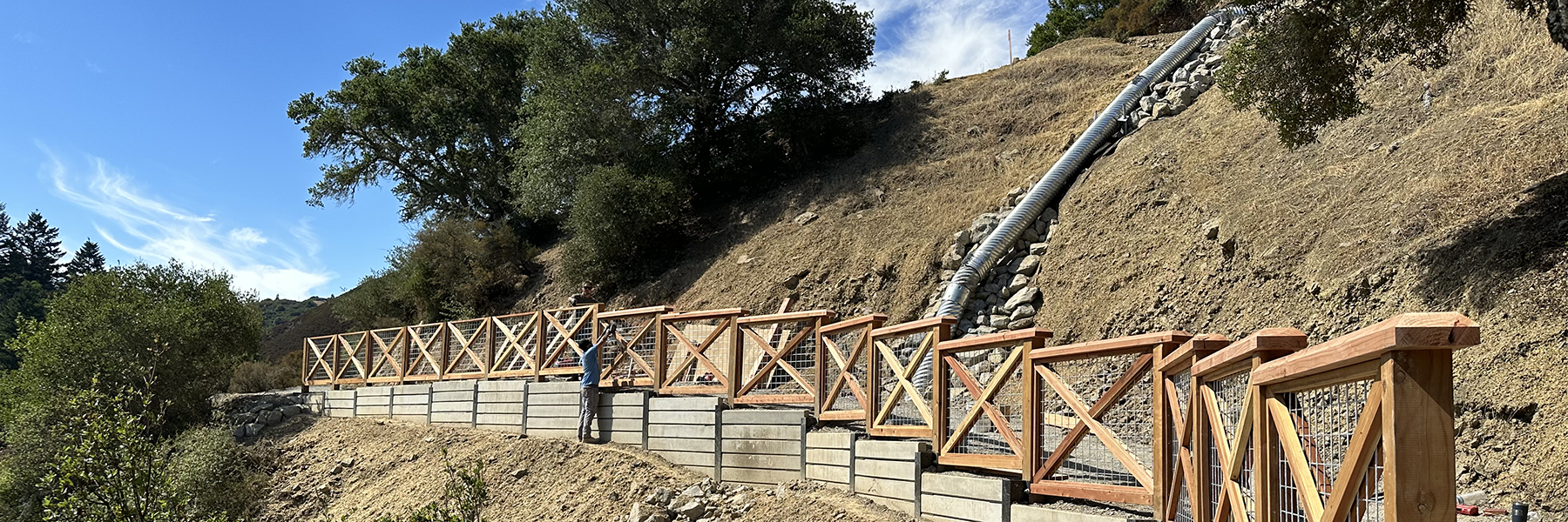 A man reaches toward a fence on a retaining wall along a hillside. A culvert runs down the hillside.