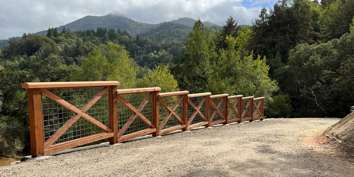 Fencing lines a dirt roadway overlooking misty, tree-covered mountains.