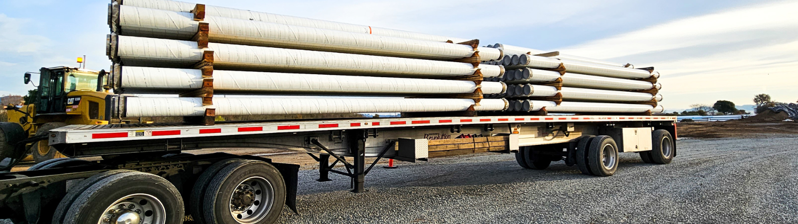 White pipes are stacked on a semi-tractor trailer in a gravel lot.