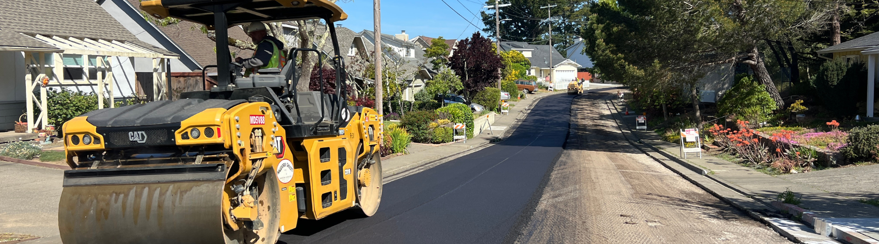 A steam roller rolls over freshly poured asphalt on a residential roadway.