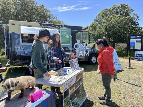 Watershed ambassadors stand behind a table, with the One Tam van in the background, and share information with a visitor. 
