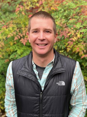 A man posed for a photo in front of a leafy background