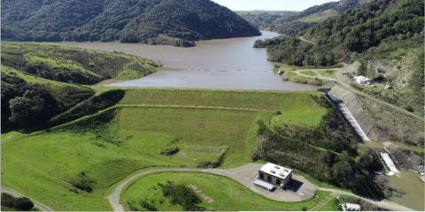 Aerial photo shows a body of water held back by an earthen dam, with spillway along the right side. 