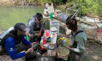 Three people use fish counting equipment on the shores of a creek. Two people in the background check the smolt trap equipment on the creek.