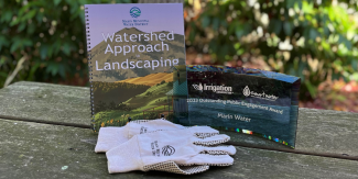 A display on an outdoor table of the Watershed Approach to Landscaping guidebook, a semi-transparent award and gardening gloves