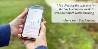 A person holds a phone displaying a water-saving app. A grey box with blue text is superimposed above, reading, "“I like checking the app, and I’m starting to compare week-to-week how much water I’m using.”   - Anne, from San Anselmo."