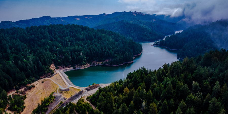 A reservoir is held back by an earthen dam and surrounded by forested hillsides.